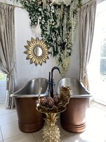A bathroom with two large copper sinks, a gold pineapple-shaped table, and a decorative mirror with a sun design.