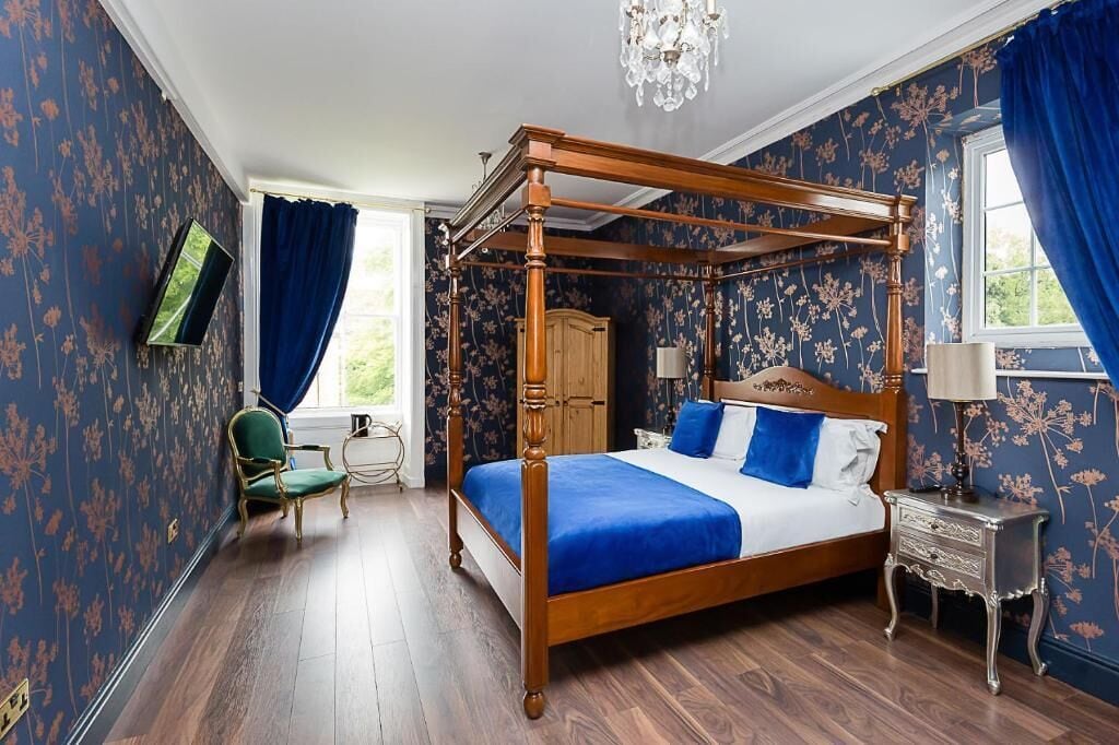 A four-poster bed with a blue bedspread, a wooden nightstand, a green armchair, and a television on the wall.