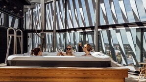 Un jacuzzi en la azotea con tres personas, ventanas amplias y vista panorámica de la ciudad.