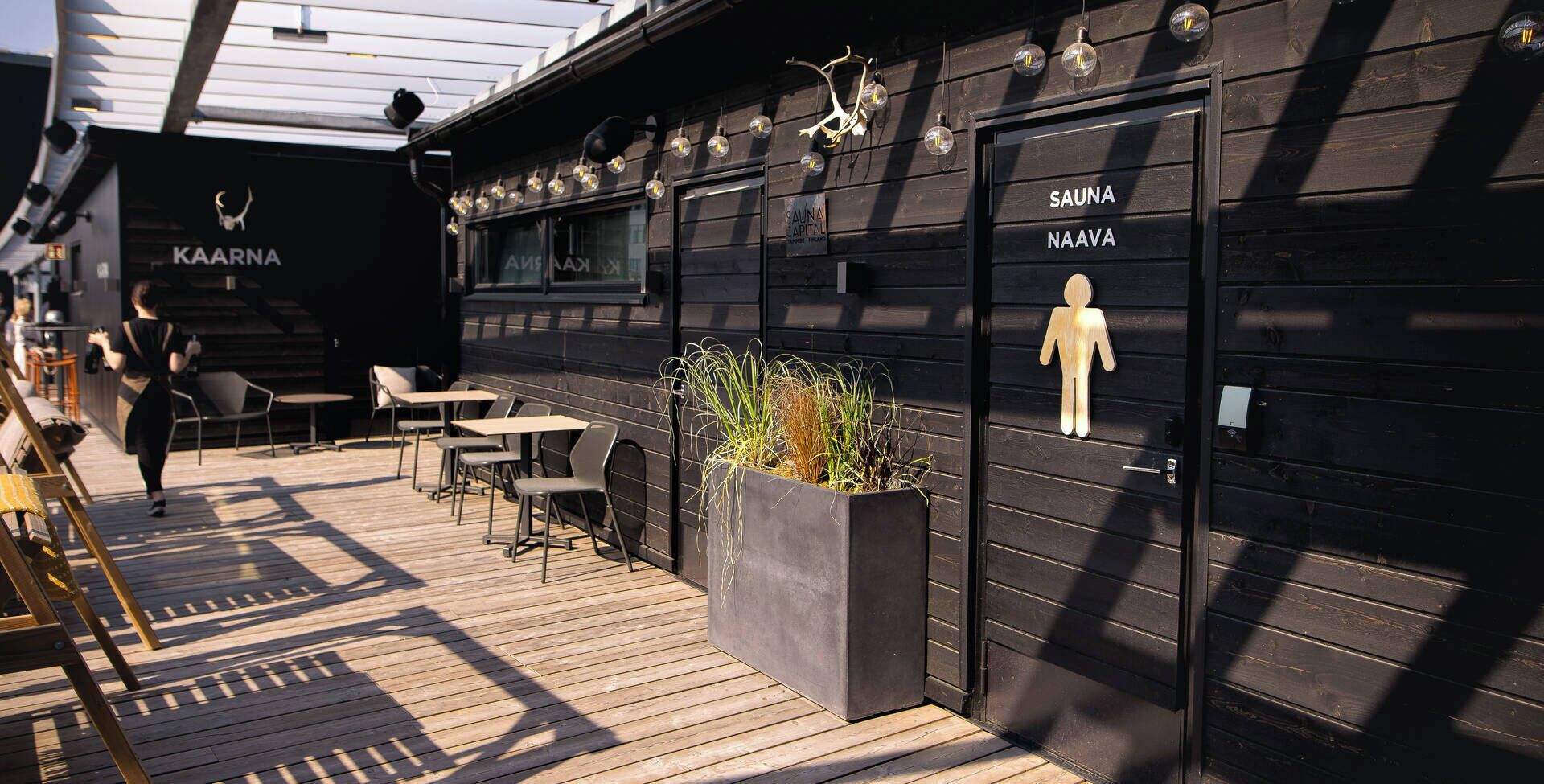 Puisesta materiaalista rakennettu terassi, jossa on ulkotilaa istumiseen, sauna ja kyltti, jossa lukee ”SAUNA NAAVA”.