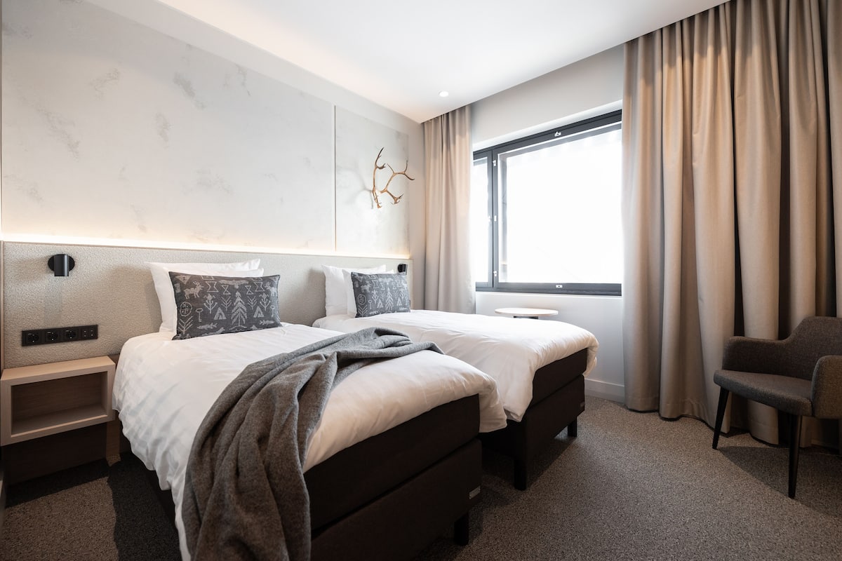 Ein modernes Hotelzimmer mit einem großen Bett, Nachttischen, einem Sessel und einem Fenster mit Vorhängen.