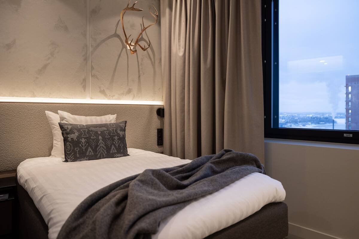 Ein modernes Hotelzimmer mit einem ordentlich bezogenen Bett, einem großen Fenster mit Blick auf die Stadt und einem dekorativen Wandbild.