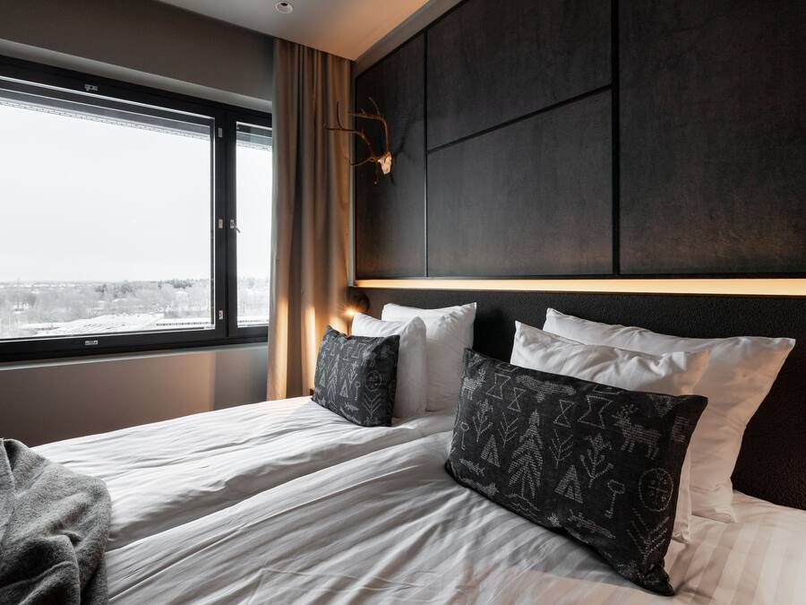 Una habitación de hotel con una cama grande, dos almohadas, una colcha, una ventana con vistas y una pared oscura con una lámpara.