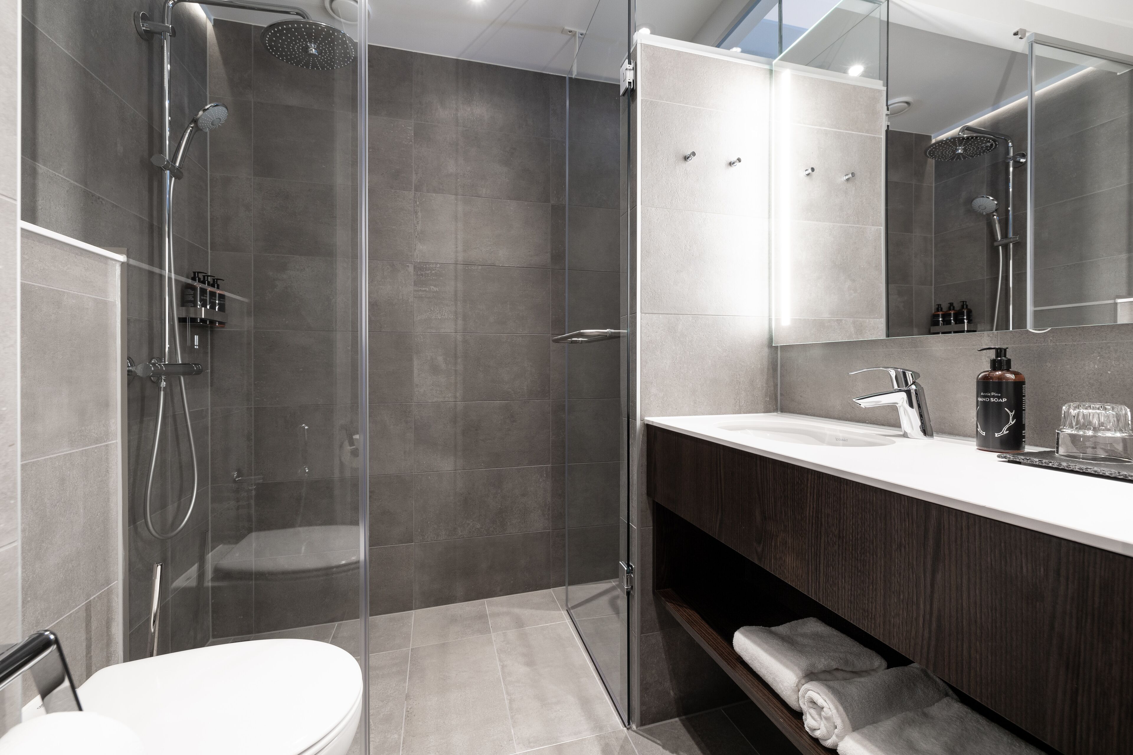 A modern bathroom with a glass-enclosed shower, a white toilet, a dark wood vanity, and a large mirror.