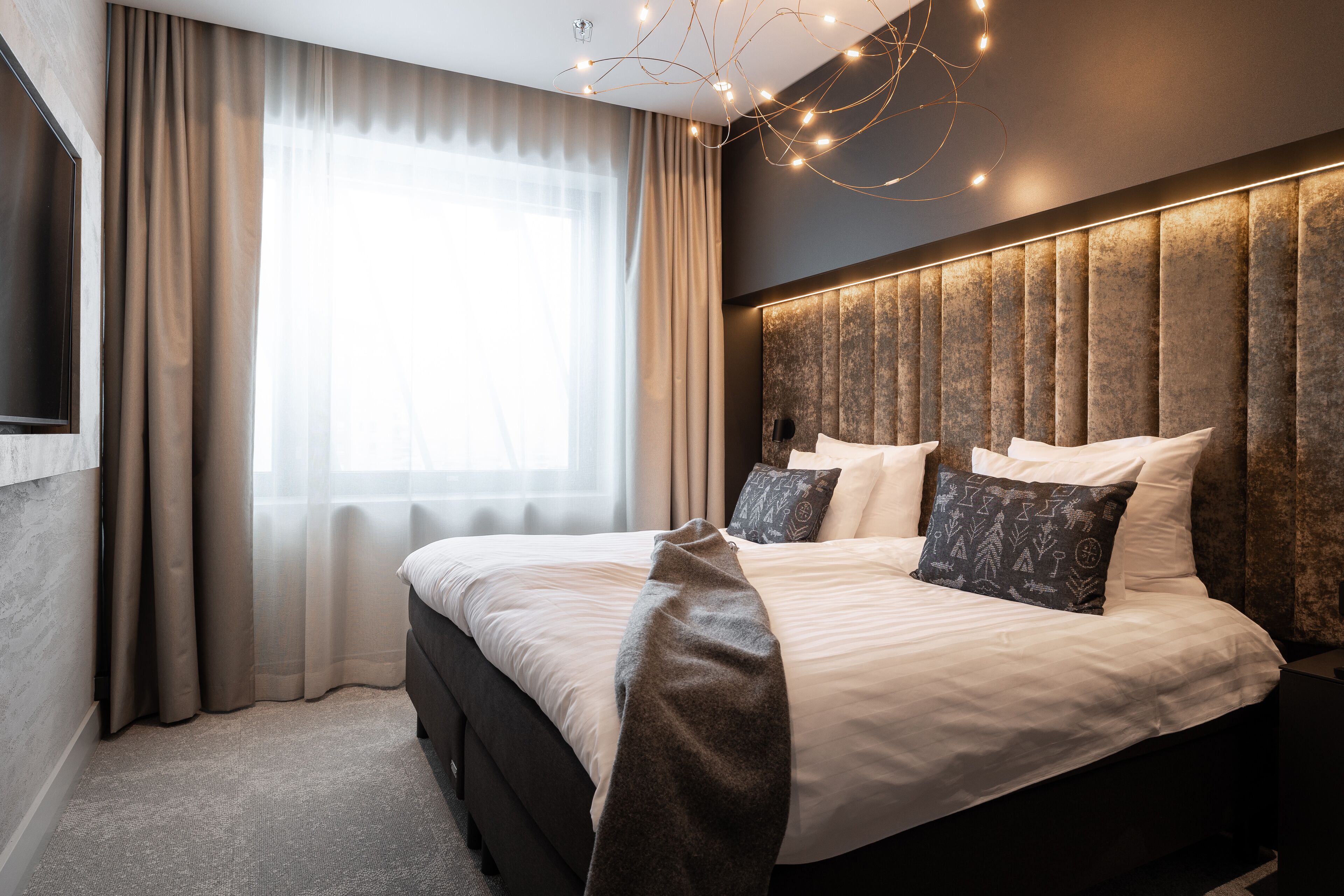 Une chambre à coucher moderne avec un grand lit, une tête de lit ornée de panneaux verticaux et une grande fenêtre avec des rideaux légers.
