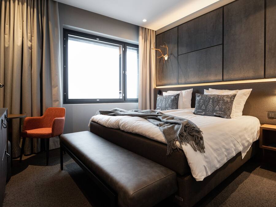 A modern hotel room with a large bed, a bench, a desk, and a window with curtains.
