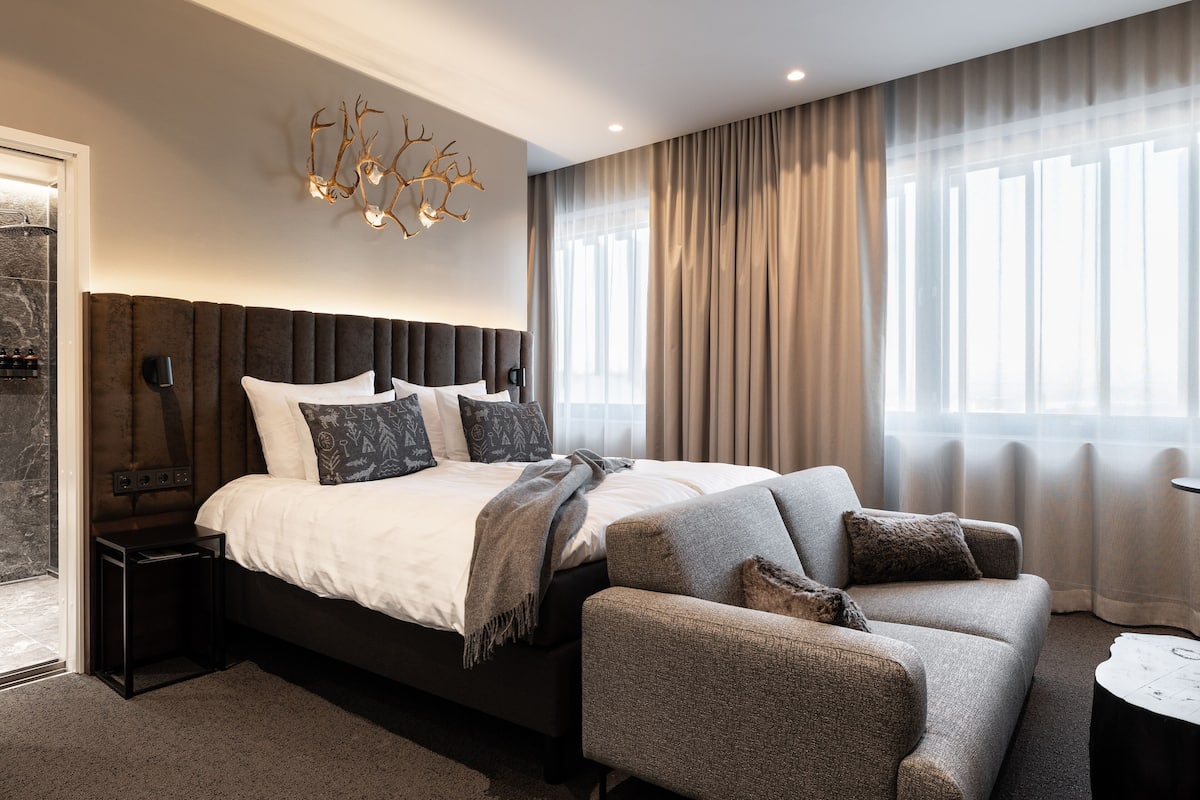 Ein modernes Schlafzimmer mit einem großen Bett, einem Kopfteil mit integrierter Beleuchtung, einem Nachttisch, einer Couch und Blick ins Freie durch transparente Vorhänge.