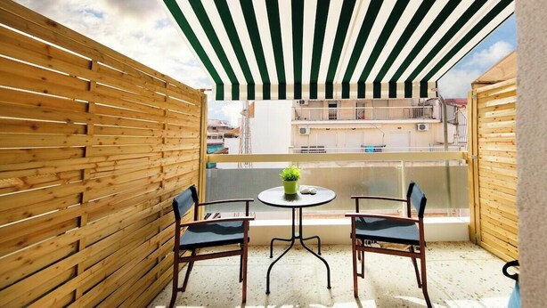 A balcony with a striped awning, two chairs, a small table with a plant, and a view of buildings. - Dimitsanas25-Startupper's Pad in real Athenian nei (Athens)