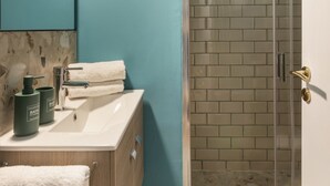 Ein Badezimmer mit einem weißen Waschbecken, einem Spiegel und einem Handtuch, das an der Tür hängt. Es gibt eine Dusche mit Glastür und einer gefliesten Wand.
