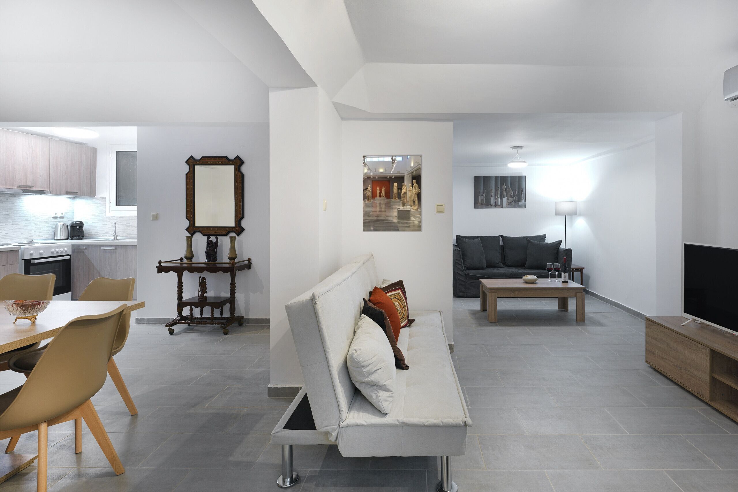A modern living room with a sofa, coffee table, and TV stand. There is a dining area with a table and chairs. The room has a large mirror and artwork on the walls.