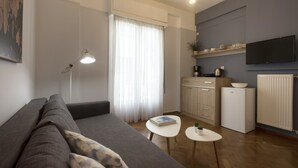Een moderne woonkamer met een grijze bank, een klein wit tafeltje en een keuken met kasten en een koelkast.