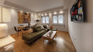 A modern living room with a sofa, coffee table, and a dining area with chairs and a table. There is a wall-mounted TV displaying a movie poster. - Karneadou34 · Stylish Kolonaki 2 Bdr apartment (Athens)