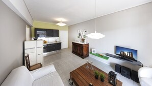 Uma sala de estar moderna com um sofá, mesa de centro e televisão. Cozinha visível ao fundo, com televisão e armários.