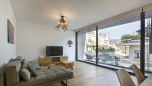 Un salon moderne avec un canapé, un téléviseur, des tables d’appoint en bois et une grande fenêtre donnant sur la ville.