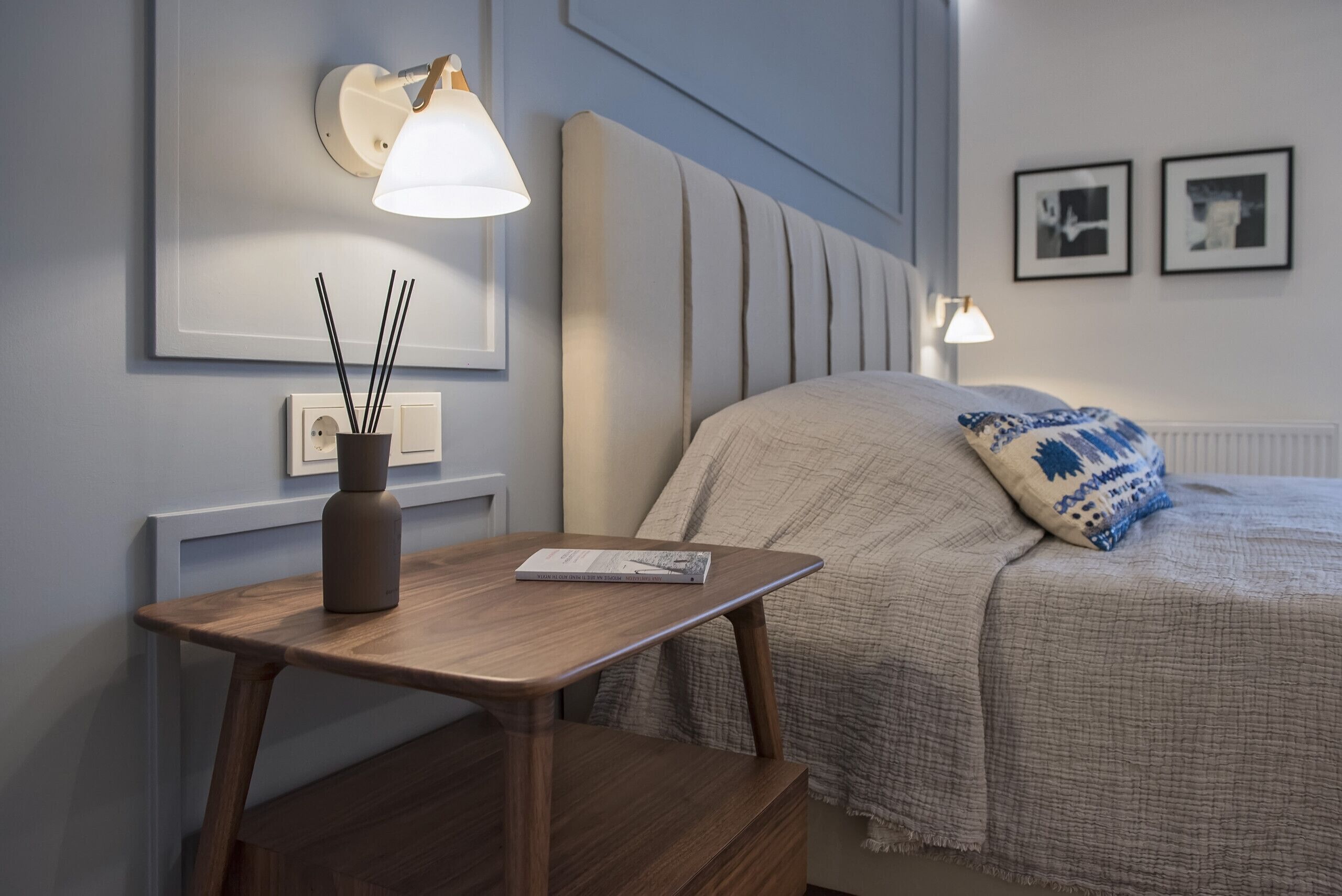 Une chambre à coucher bien rangée, avec un lit, des lampes de chevet, une table de chevet sur laquelle repose un livre, et un vase contenant des tiges de roseau.