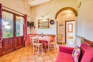 A dining area with a table set for two, a television on the wall, and a view of the garden through the window.
