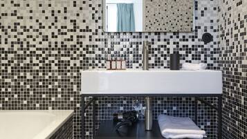Een badkamer met een betegelde mozaïekwand, een wit wastafelmeubel, een zwart plankje en een spiegel boven de wastafel.