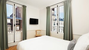 A hotel room with a large bed, a mounted TV, and a view of buildings through the windows. - My Maison In Paris - Champ de Mars (Paris)