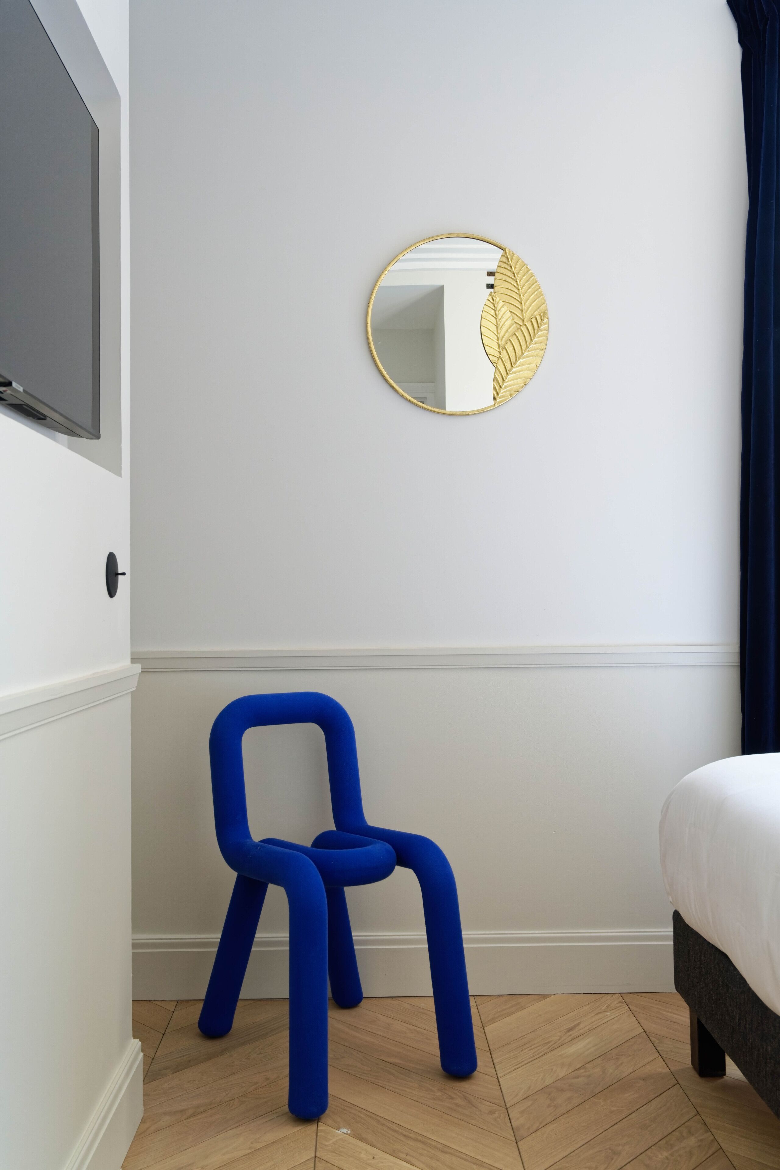 A room with a bed, a blue chair, a round mirror with a leaf design, and a television on the wall.