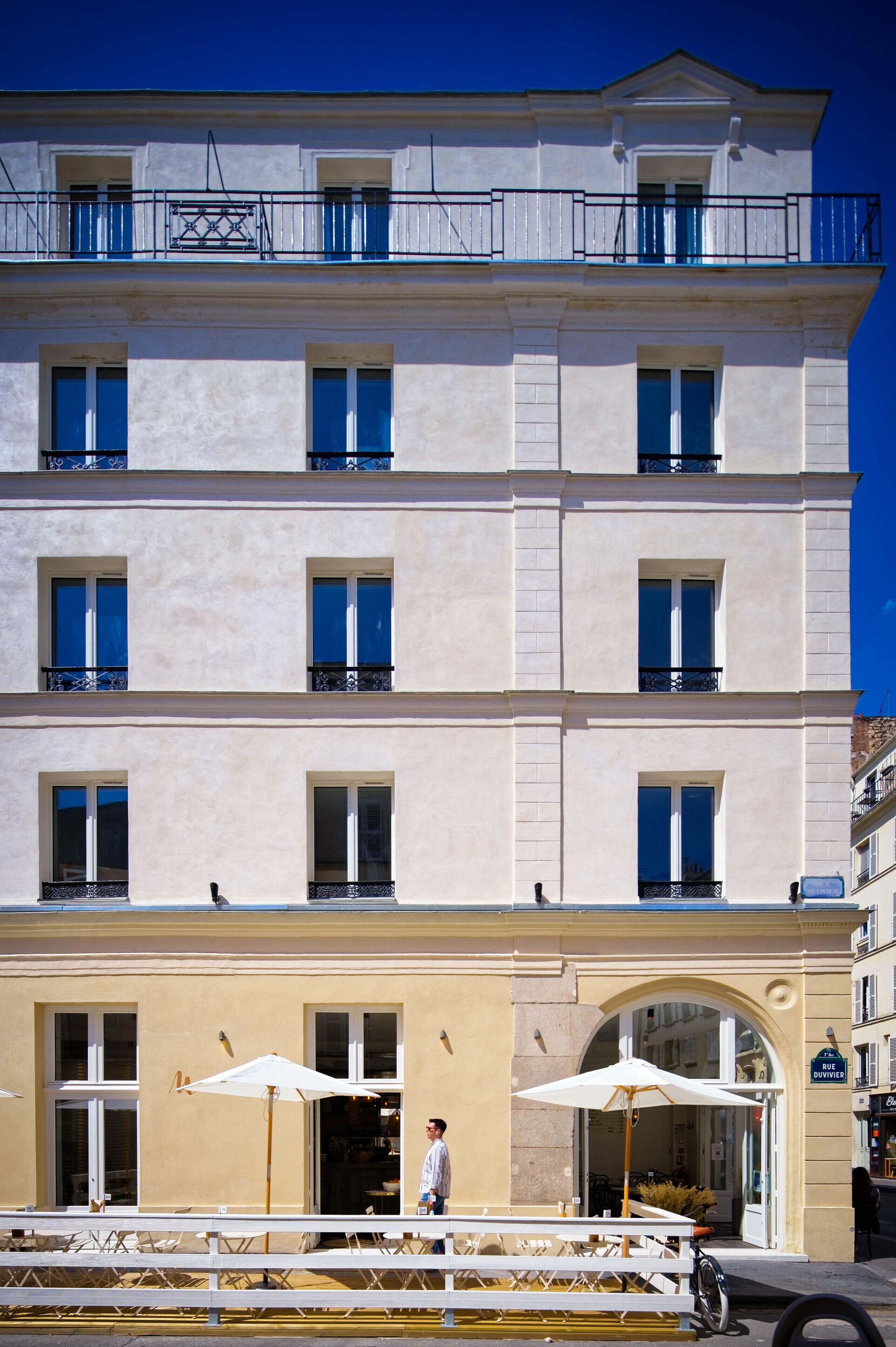 A multi-story building with balconies, a ground-level entrance with an archway, and outdoor seating with umbrellas.