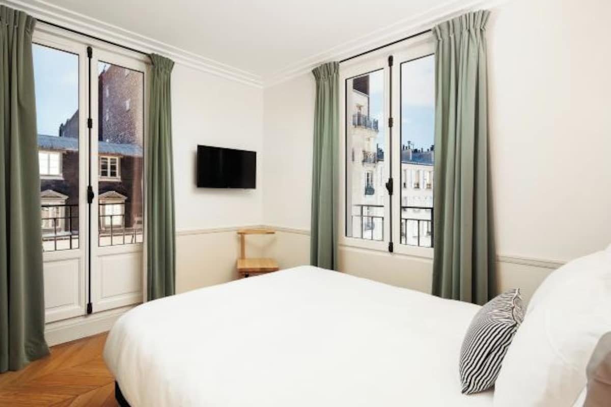Une chambre d’hôtel avec un grand lit, un téléviseur fixé au mur et une vue sur les immeubles par les fenêtres.