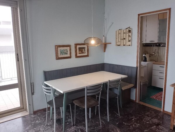 Un comedor con una mesa y sillas, una lámpara colgante, cuadros en la pared y una cocina visible a través de un umbral abierto.