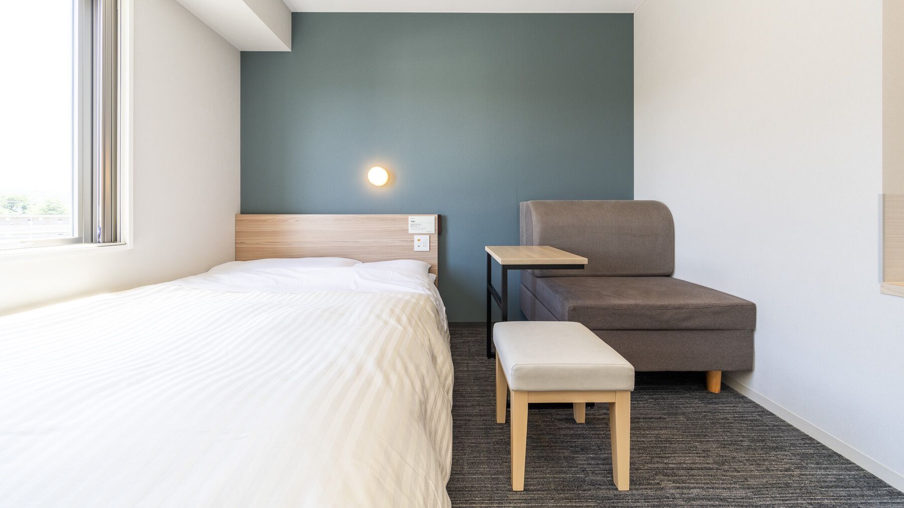 Une chambre d’hôtel avec un lit, une chaise, une petite table et une lampe fixée au mur.