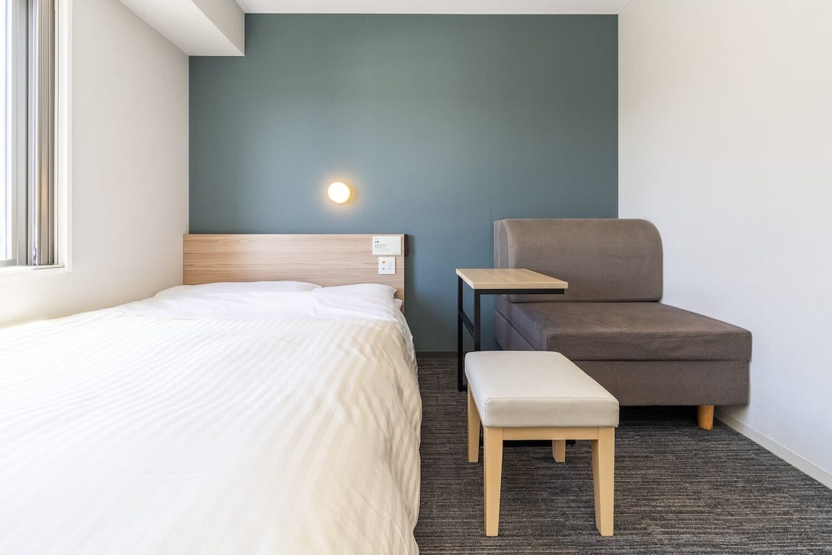 Een hotelkamer met een bed, een stoel, een klein tafeltje en een wandlamp.
