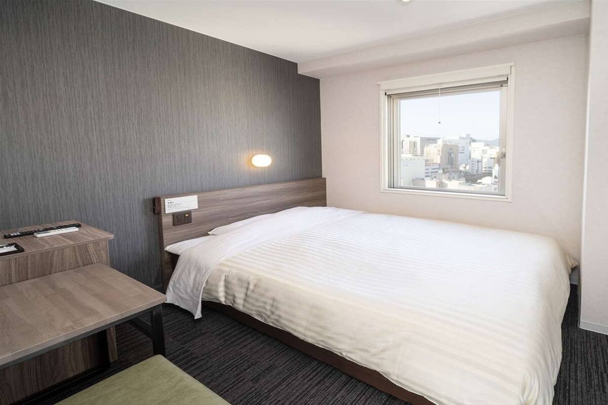 Hotelkamer met een bed, bureau, stoel, nachtkastje, raam met uitzicht op de stad en een wandlamp.