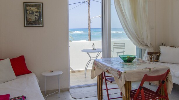 Una habitación con una cama, una mesa con un cuenco verde, una silla roja y dos ventanas con vistas a la playa.
