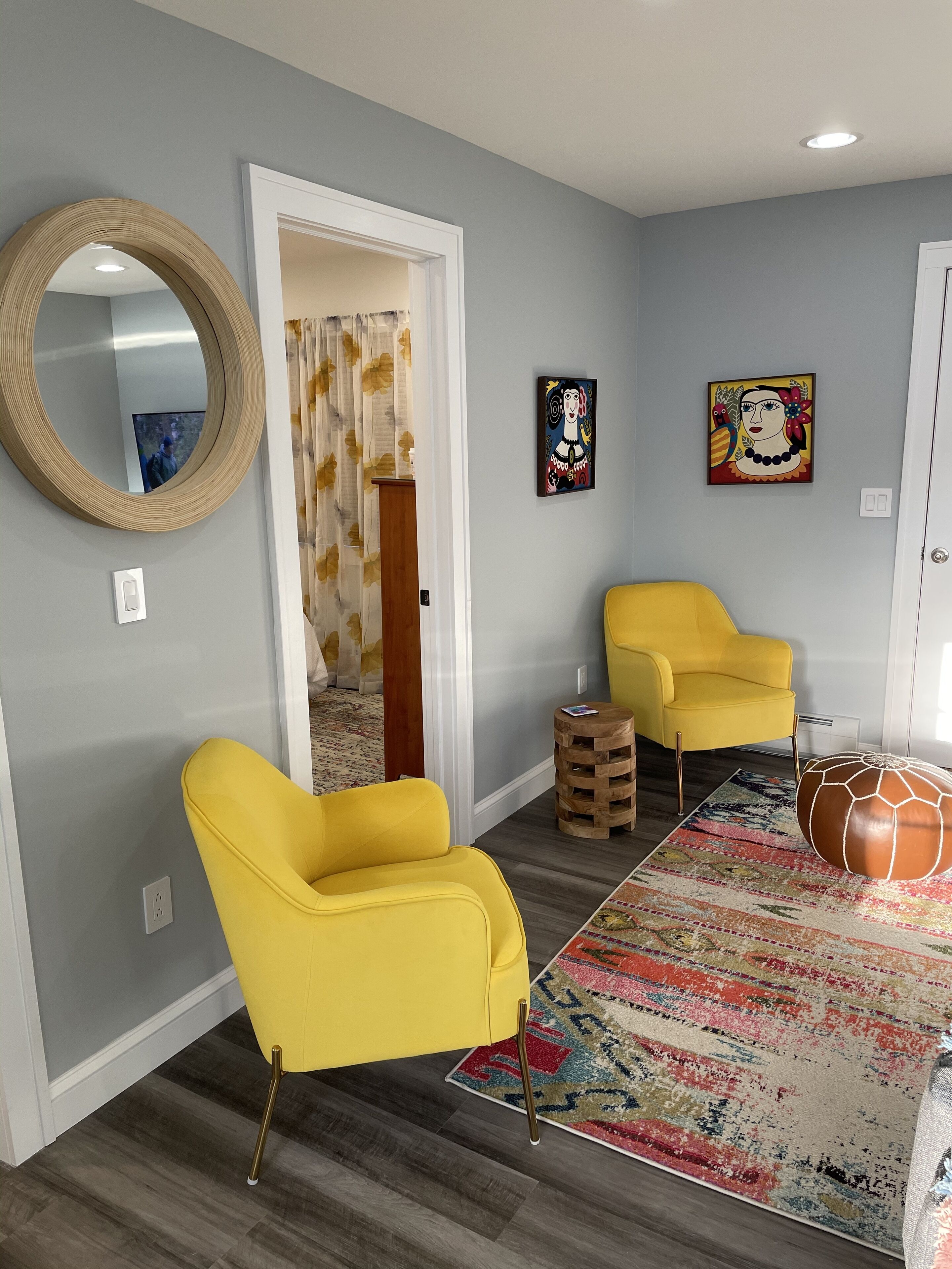 A room with two yellow armchairs, a round mirror, a colorful rug, and a wooden stool.