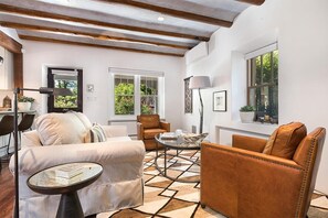 A living room with a sofa, armchair, coffee table, and a patterned rug. There are windows with views of greenery, a lamp, and a mirror.