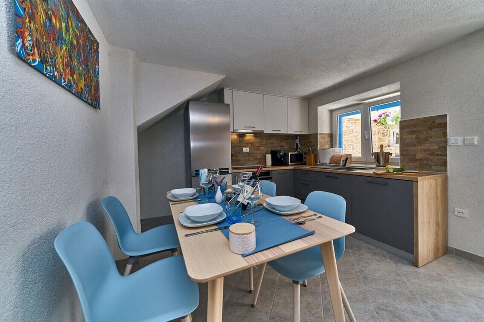 A modern kitchen with a dining area, featuring a large painting, contemporary appliances, and a view to the outside.