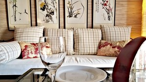A modern living room with a glass coffee table, a wine glass, and a white plate. There is a sofa with striped cushions and floral pillows. Behind the sofa, there are framed artworks on the wall.