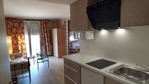 A modern kitchen with a microwave, stove, and sink. There is a dining area with a table and chairs. The room has wooden flooring and a large window with curtains.
