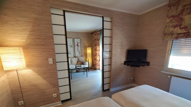A hotel room with a bed, a TV, a lamp, and a view of another room with a sofa and a window.