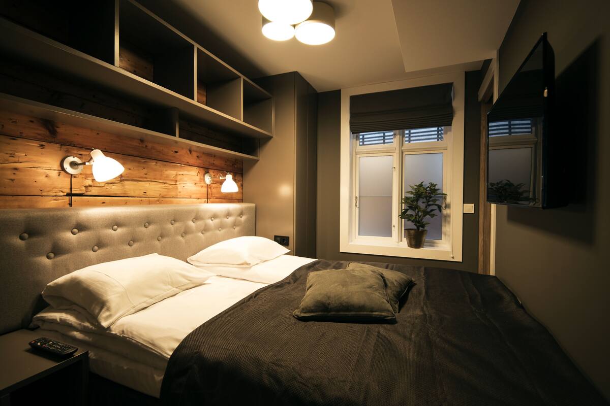 A bedroom with a tufted headboard, a bed with white and dark bedding, a bedside table with a remote control, and a window with a plant.