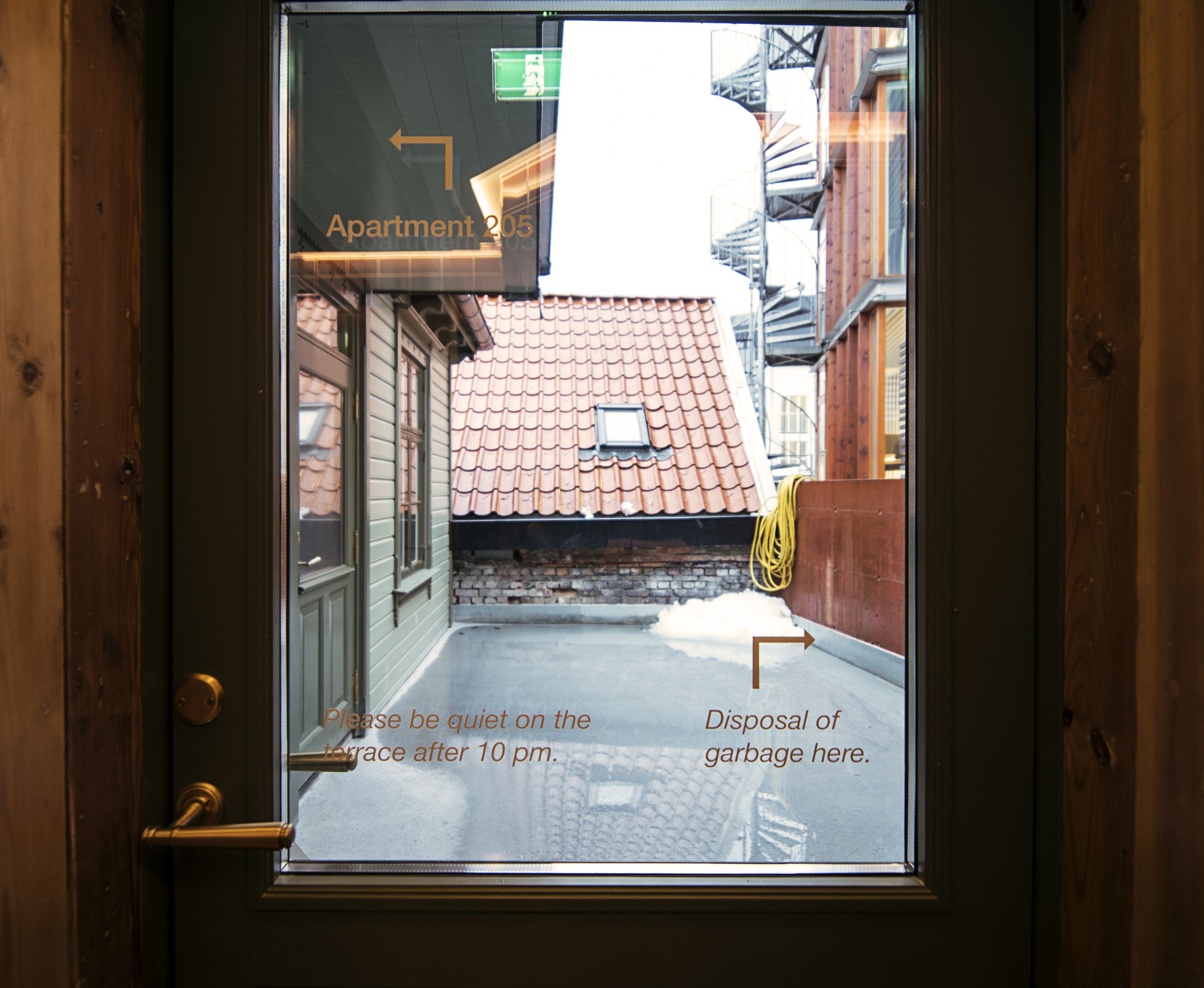 A view through a window of a building with a tiled roof, a sign indicating 'Apartment 205', and a notice about quiet hours and garbage disposal.