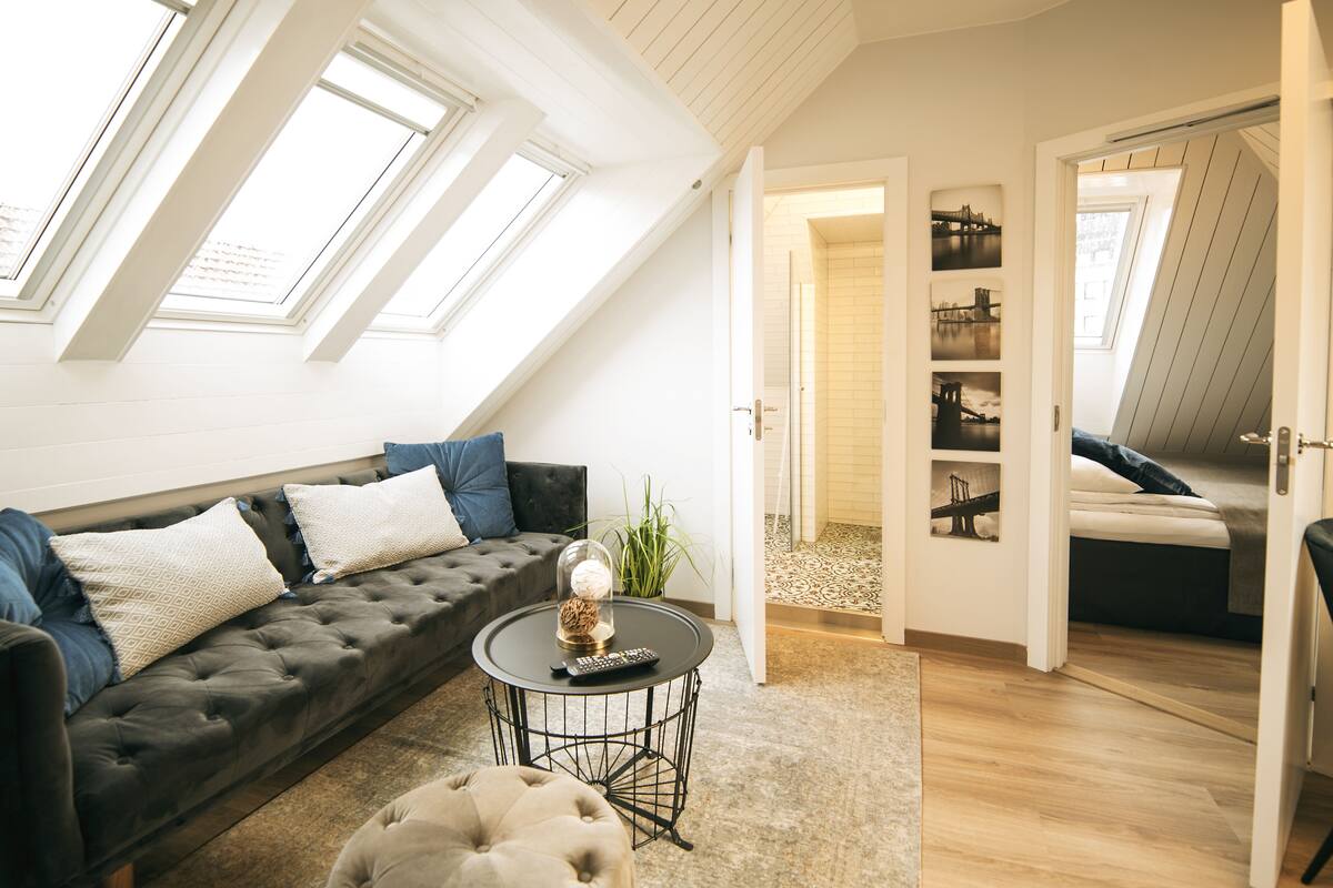 A loft-style room with a dark grey sofa, a round coffee table, and a small ottoman. There are skylights and a view of a cityscape through the open door.