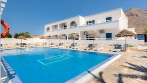 A swimming pool with lounge chairs and umbrellas, a white building with multiple balconies, and a clear blue sky.