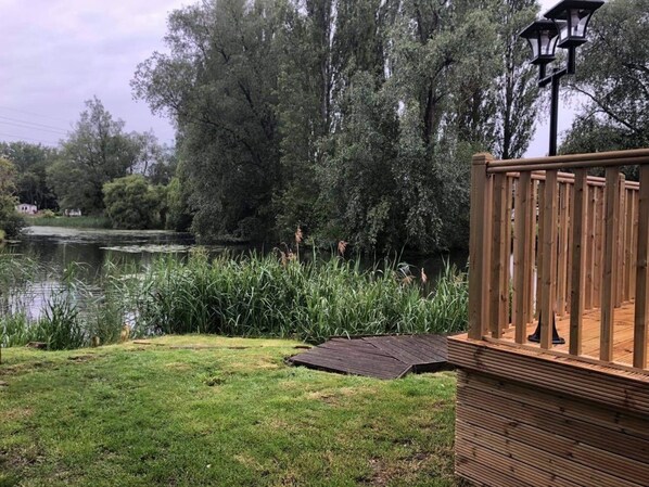 A wooden deck with a metal railing, a pond, and dense trees in the background. - Beautiful and Tranquil Lakeside Lodge - Sleeps 5 (Northampton)