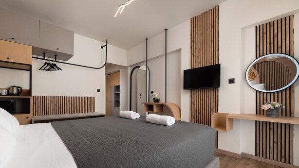 A modern hotel room with a large bed, wooden paneling, a flat-screen TV, a round mirror, and a small shelf with a plant. - Santa Maria City Hotel (Rhodes)