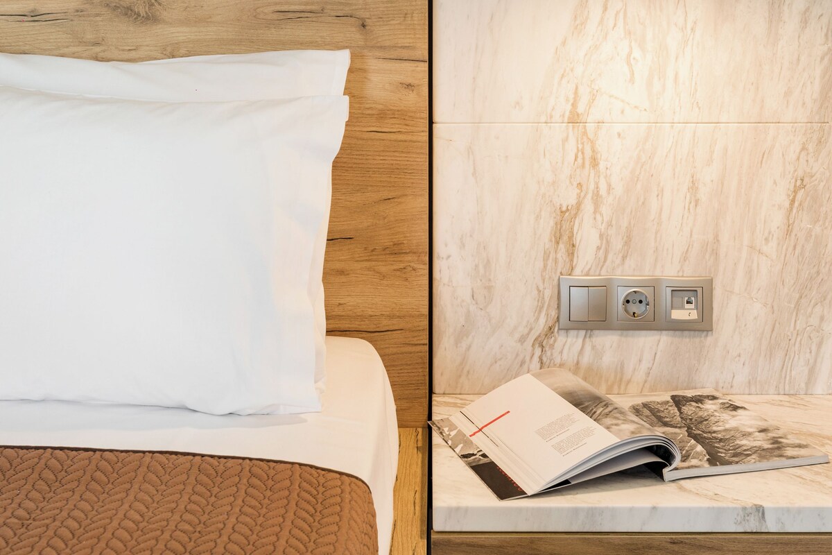 A bed with a white pillow and a brown blanket, a wooden headboard, a wall-mounted reading light, and a magazine on a nightstand.