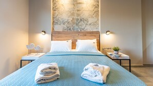 A modern bedroom with a wooden headboard, two bedside tables with lamps, a blue quilted bedspread, and neatly folded towels. - Olympus Pantheon Luxury Rooms (Dio-Olympos)