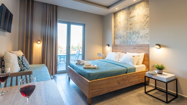 A modern hotel room with a wooden bed, a small table with glasses of red wine, a view through a sliding glass door, and a wall-mounted TV. - Olympus Pantheon Luxury Rooms (Dio-Olympos)