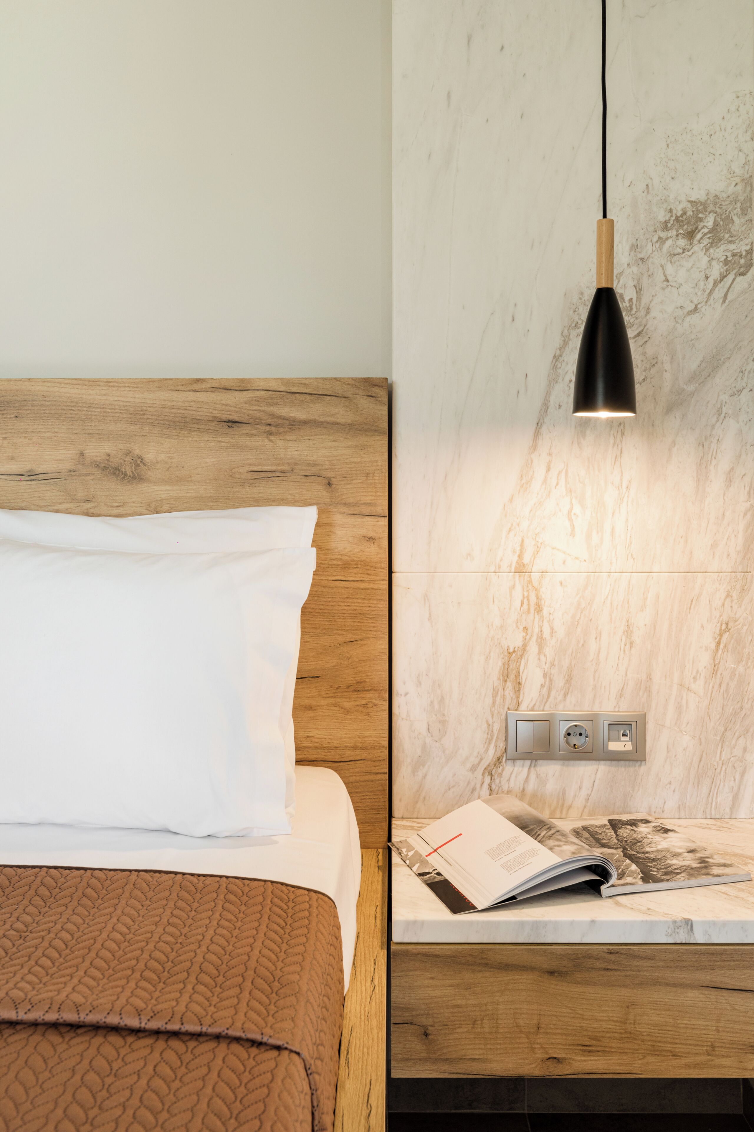 A bed with a white pillow and a brown blanket, a wooden headboard, a wall-mounted reading light, and a magazine on a nightstand.