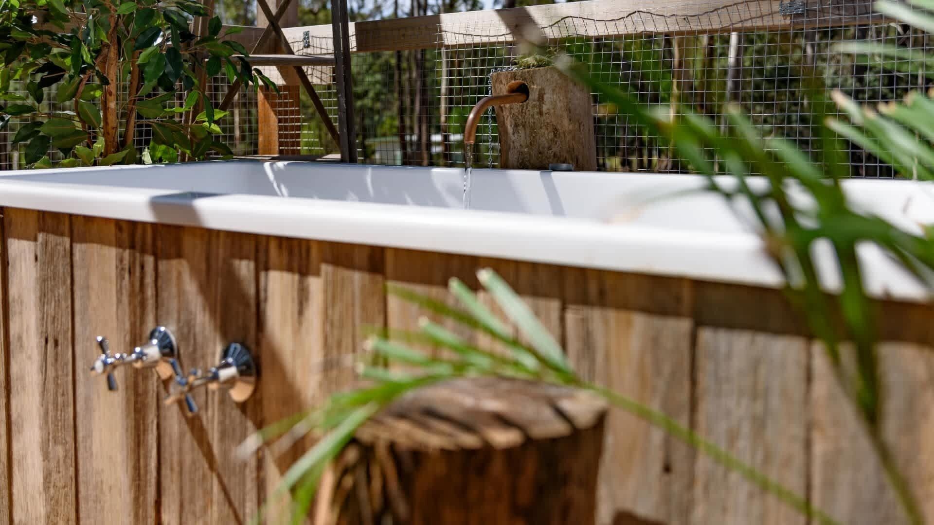 Une terrasse en bois avec une baignoire blanche, un robinet en métal et un tabouret en bois.