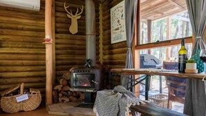 A cozy cabin interior with a wood-burning stove, a log storage area, and a window with a view of the forest. - The Love Shack (Wheatsheaf)