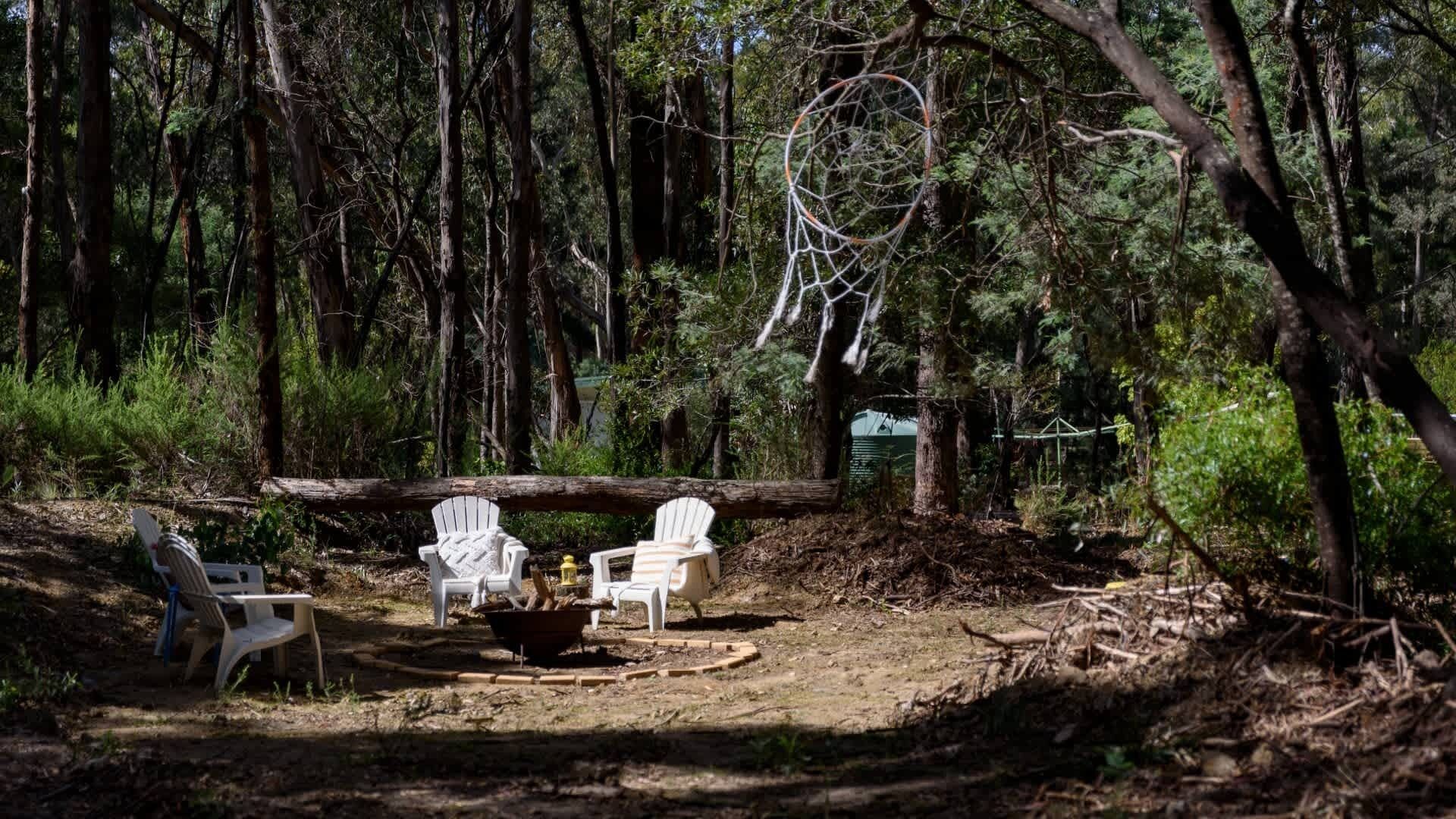 Un claro en el bosque con dos sillas blancas, una fogata y un atrapasueños colgado de un árbol.