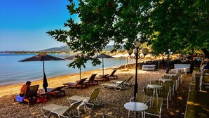 Un café frente a la playa con terraza, que incluye tumbonas y mesas, con vista a una masa de agua tranquila y una montaña distante.